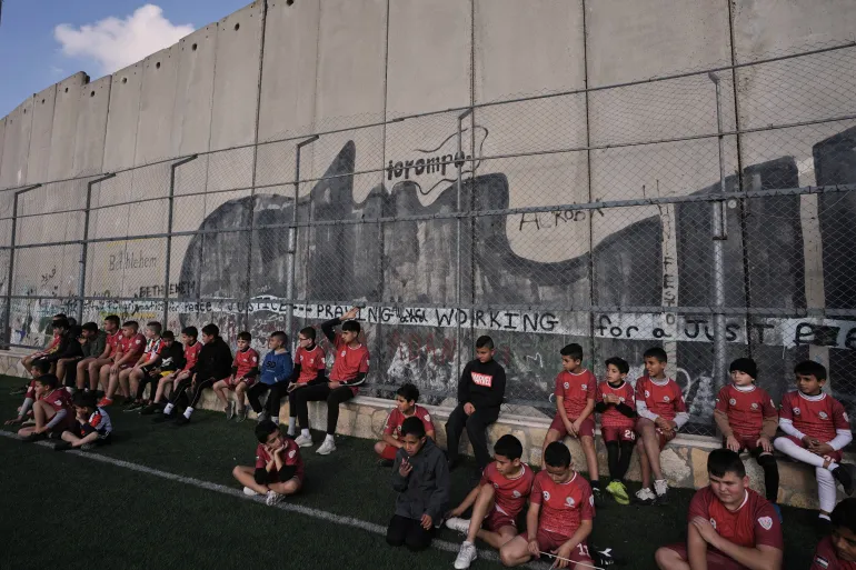 Giovani palestinesi partecipano a una sessione di allenamento di calcio in un campo vicino al muro di separazione israeliano nel campo profughi di Aida, nella città di Betlemme, in Cisgiordania, venerdì 26 dicembre 2025, settimane dopo che le autorità israeliane hanno emesso la decisione di demolire il campo. (AP Photo/Mahmoud Illean)
