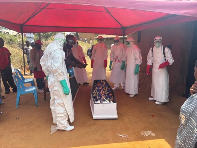 Una squadra di sepoltura della Croce Rossa in equipaggiamento protettivo prepara una bara sotto gli occhi dei parenti, parte di un processo altamente controllato progettato per prevenire un’ulteriore trasmissione di Ebola.